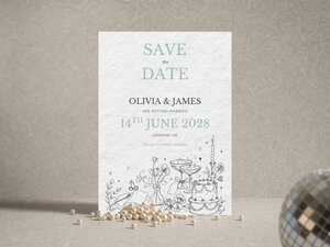A save-the-date card with travel-themed line art and text for "Olivia & James" stands upright on a gray surface with scattered pearls.