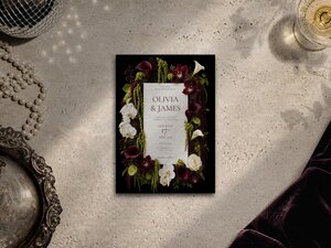 A wedding invitation card with dark floral border of burgundy calla lilies and white orchids rests on a textured stone surface next to pearls and velvet fabric.
