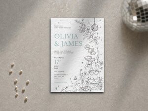 A wedding invitation with a blue and white floral design lies flat on a gray surface, with a disco ball and scattered pearls nearby.