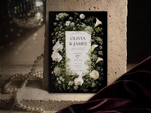 A black wedding invitation featuring white flowers like orchids and roses rests against a rustic stone and pearl background.