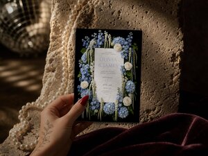 A hand with red nail polish and a floral tattoo holds the wedding invitation against a textured surface with pearls and velvet fabric.