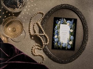 The wedding invitation rests inside an ornate round silver tray on a textured tabletop alongside a string of pearls and a glass of champagne.