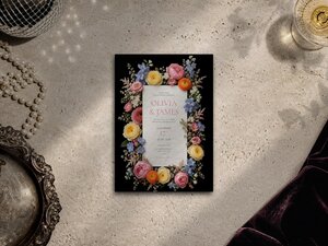 A flatlay view of a floral wedding invitation on a rustic stone surface beside pearls, a silver tray, and dark fabric.