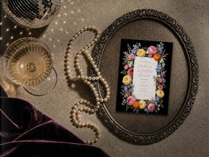 A flatlay shows a floral wedding invitation inside an ornate oval metal tray next to a string of pearls, a glass of champagne, and velvet cloth.