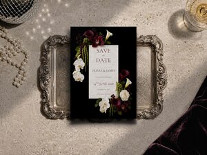 A Save the Date card featuring dark burgundy and white flowers rests on a detailed silver tray on a textured background.