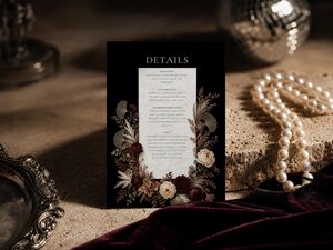 An upright details card for a wedding stands on a stone ledge next to a strand of pearls and dark velvet fabric, with a disco ball reflecting light in the background.