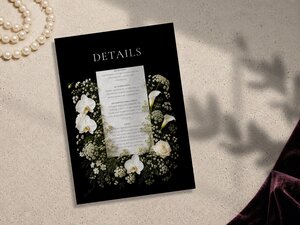 A black details card adorned with white orchids and floral elements sits flat on a textured surface.