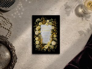 Flat lay of a black wedding invitation template with yellow flowers on a textured stone background alongside pearls, a silver tray, and a glass of champagne.