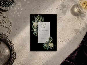 A black save the date card with sage green floral corners and white text on a neutral textured background.