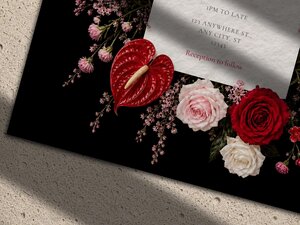 A close-up view of the bottom corner of a black wedding invitation showcasing red and pink roses alongside an anthurium flower.