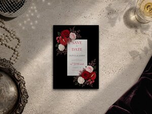 A black save the date card featuring red and white floral arrangements is placed on a textured surface.