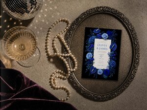 A single wedding invitation card with blue florals rests inside an ornate silver tray next to pearls and a glass of champagne.