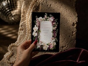 A hand with red nail polish and a floral tattoo holds the wedding invitation card in front of a textured stone block with pearls and a disco ball in the background.