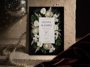 A vertical wedding invitation template featuring tropical white florals and dark foliage stands upright against a stone surface.