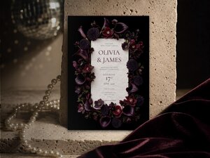 Plum floral wedding invitation featuring plum calla lilies, dark dahlias, burgundy orchids and velvety hellebore flowers arranged around elegant wedding stationery on a dramatic black background.