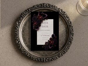 Plum floral wedding save the date featuring plum calla lilies, dark dahlias, burgundy orchids and velvety hellebore flowers arranged around elegant wedding stationery on a dramatic black background.