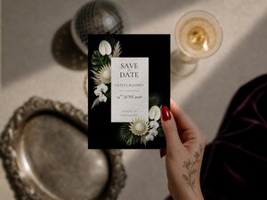 A hand with red nails and a floral wrist tattoo holds a save-the-date card template against a neutral background with decorative props.