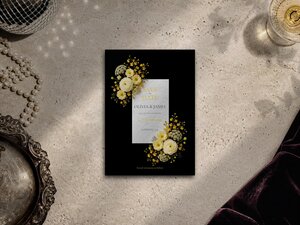 A Save the Date card with yellow and white flowers on a black background lies flat on a textured surface alongside a tray and pearls.