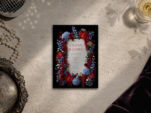 A wedding invitation template featuring dark floral borders with red and blue flowers lies on a textured surface next to pearls and a silver tray.