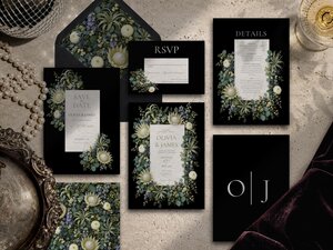 A flat lay of a complete black wedding invitation suite with sage green floral illustrations, pearls, and a glass of champagne.