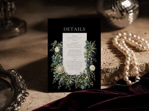 A black wedding details card with a white text block framed by sage green florals resting on a textured surface.