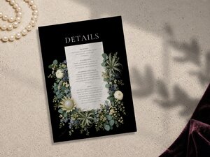 A black wedding details card featuring a white central text block surrounded by sage green florals and white protea flowers, resting on a textured surface next to pearls.