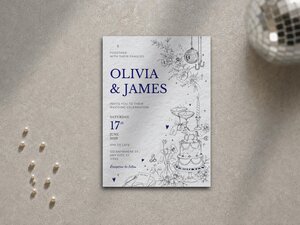 A wedding invitation card styled with blue text and line drawings resting on a grey textured surface next to scattered pearls and a disco ball.