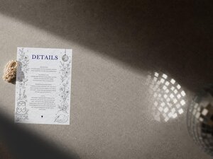 A wedding details card with hand-drawn illustrations placed on a textured grey surface beside a bracelet of pearls and a disco ball casting light reflections.