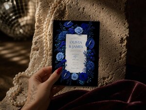 A hand holds a vertical wedding invitation card featuring blue roses and anthuriums against a black background.