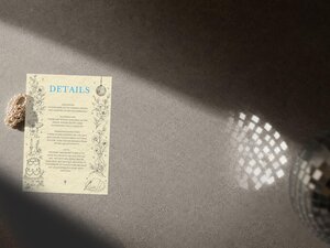 A wedding details card with blue typography and floral line art resting on a textured gray surface beside pearls.