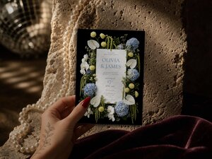 A hand holds a wedding invitation card featuring blue and yellow floral arrangements against a black background.