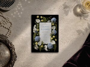 Flat lay of a floral wedding invitation card placed on a textured stone surface with pearls and velvet fabric.