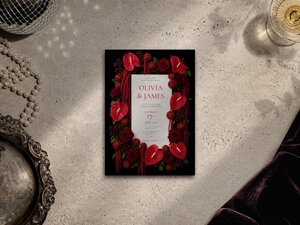 A black wedding invitation card with a border of red flowers lies flat on a textured neutral surface near a vintage silver tray and pearls.