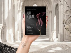 A hand holds a black wedding invitation with calla lily artwork in front of a blurred gothic architectural background.
