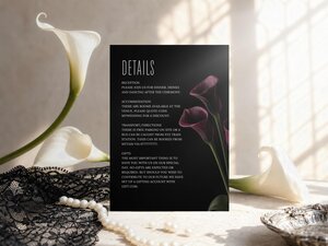 A black wedding details card with dark burgundy calla lilies is displayed upright on a table with white lilies, black lace, and pearls.