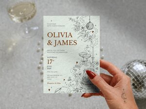 A hand holds up the light green wedding invitation featuring brown text and line illustrations.