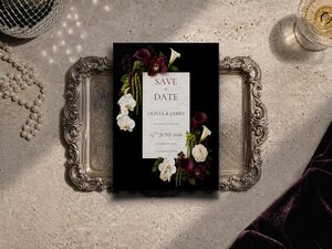 A black save the date card with burgundy and white flowers rests on a silver tray on a textured beige surface next to pearls.