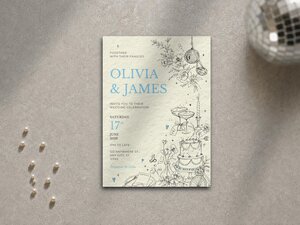 A wedding invitation card rests on a grey surface next to pearls and a disco ball with sunlight casting shadows across the scene.