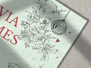 A macro shot focusing on the illustrated details of the wedding invitation, showing a T-rex dinosaur nestled among flowers and a hanging disco ball.