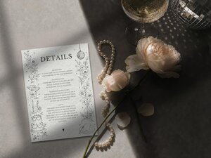 A wedding details card decorated with floral line art, paired with a strand of pearls, two pale pink roses, and a champagne glass.