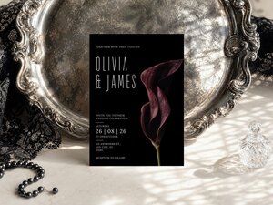 A wedding invitation with a dark burgundy calla lily graphic is displayed on a decorative silver tray next to a black lace cloth.