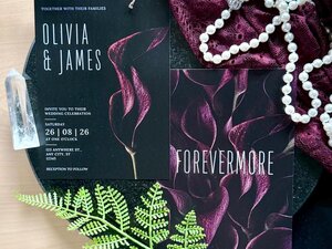 Two stationery cards with dark calla lily motifs lie on a dark surface alongside a white pearl necklace, a crystal, and a green frond.