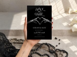 A hand holding the black save the date card with a skeletal hand heart design above a pearl necklace and tray.
