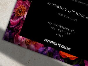 A close-up of the bottom corner of a dark floral wedding invitation showing colorful jewel-toned flowers and white text on a black background.
