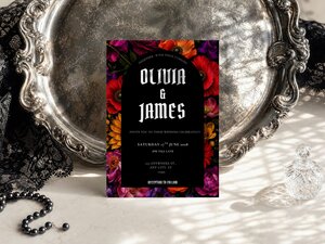A gothic floral wedding invitation centered on a ornate silver tray next to black lace and black pearl beads.