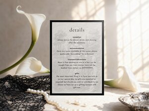 A wedding details card stands upright, flanked by white calla lily flowers, black lace, and a string of white pearls.