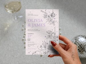 A hand holding a wedding invitation card against a grey background with a glass of champagne and a disco ball.