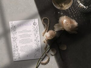 A wedding details card displayed on a grey surface alongside a pearl necklace, pale pink roses, and a glass of champagne.