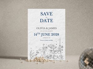 Save the date card featuring blue text and botanical illustrations, displayed on a textured surface with pearls and a partial disco ball.