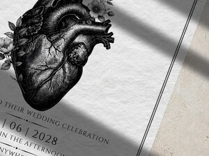 A close-up view of a wedding invitation featuring a detailed vintage botanical heart illustration and text, resting on a textured white surface.